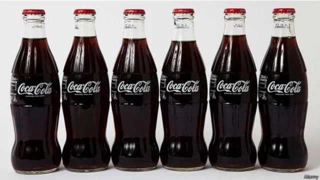 Foto de seis garrafas de vidro de Coca-Cola, idênticas, posicionadas lado a lado.