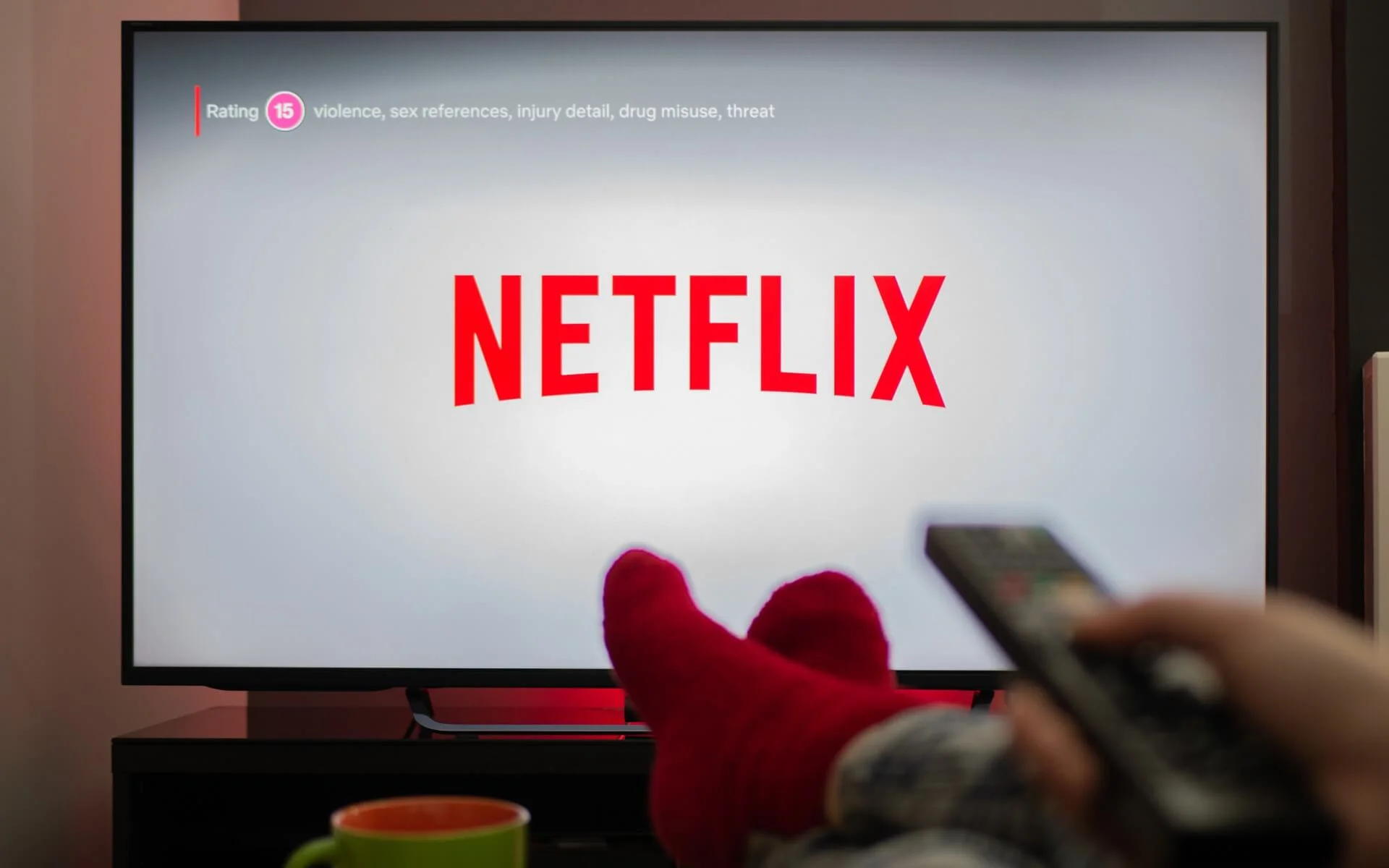 Foto de televisão exibindo o logo da Netflix. Em primeiro plano, os pés descalços de uma pessoa e sua mão segurando o controle remoto.