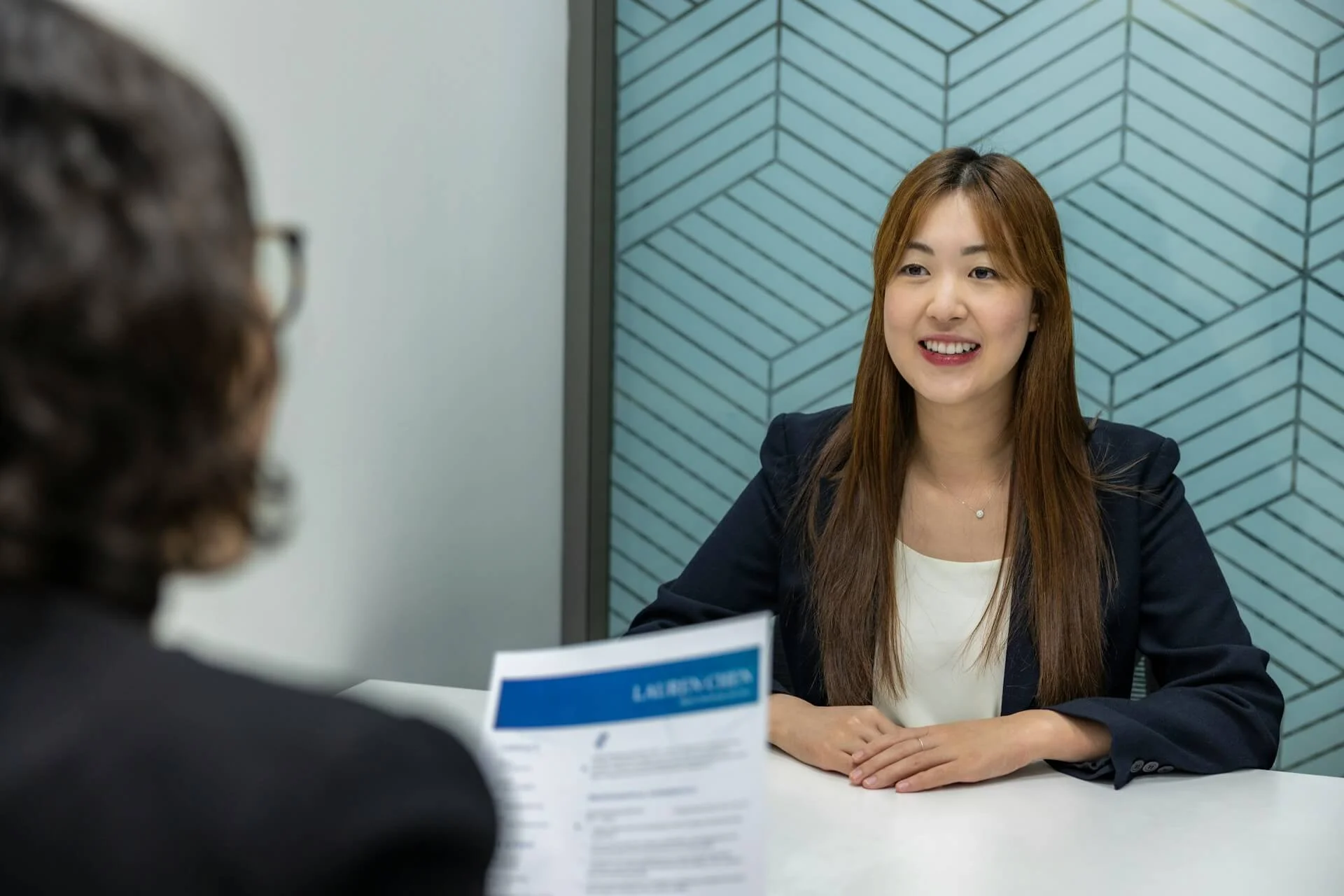 Foto de uma mulher sendo entrevistada para um emprego. A pessoa diante dela está segurando um currículo impresso.