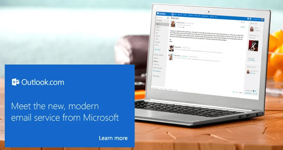 Foto de um laptop sobre uma mesa, exibindo uma tela do Outlook.com. Em uma caixa de texto azul, lê-se: “meet the new, modern email service from Microsoft”.