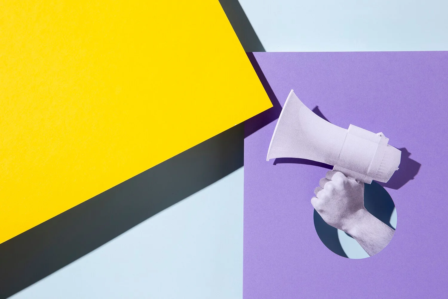 Imagem de uma mão segurando um megafone. Essa mão sai de um quadrilátero roxo, enquanto um segundo quadrilátero amarelo parece representar o som alto do megafone.