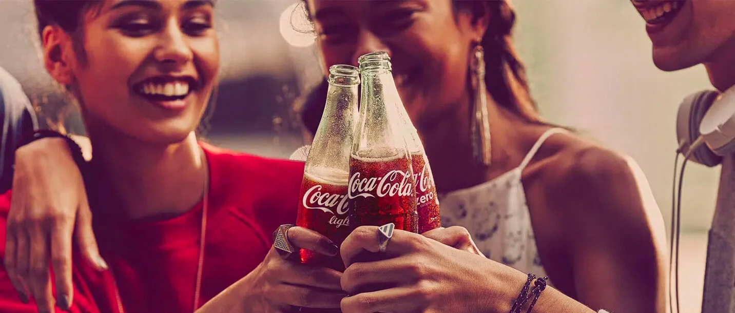 Anúncio da Coca-Cola que mostra três jovens sorridentes, brindando com suas garrafas do refrigerante.