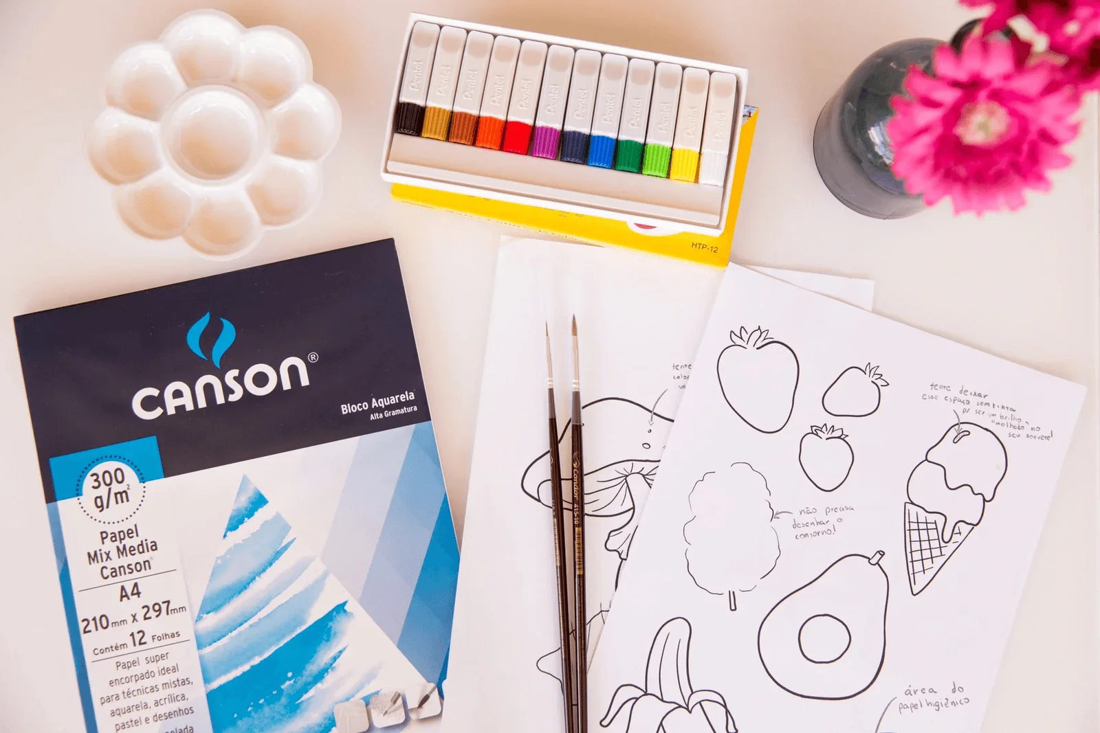 Foto de alguns itens que compõem um kit para pintar com aquarela: estojo de tintas, godê, pincéis, bloco de papel e desenhos prontos.