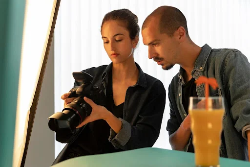 Foto de uma mulher com uma câmera fotográfica profissional nas mãos, mostrando o visor da câmera para um homem ao seu lado. Desfocado em primeiro plano, um copo de suco de laranja é iluminado por um refletor de estúdio.
