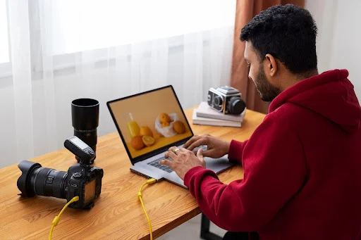 Foto de um homem sentado numa mesa, transferindo fotos de uma câmera profissional para um laptop.