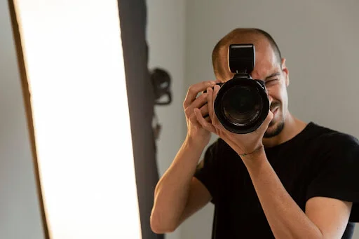 Foto de um homem olhando através do visor de uma câmera profissional, apontada para nós. Ao fundo da foto há um grande refletor.