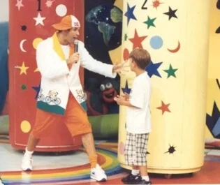 Foto antiga mostrando um homem de boné, blazer e bermuda coloridos falando com um menino diante de cabines cilíndricas fechadas, num cenário de programa infantil.