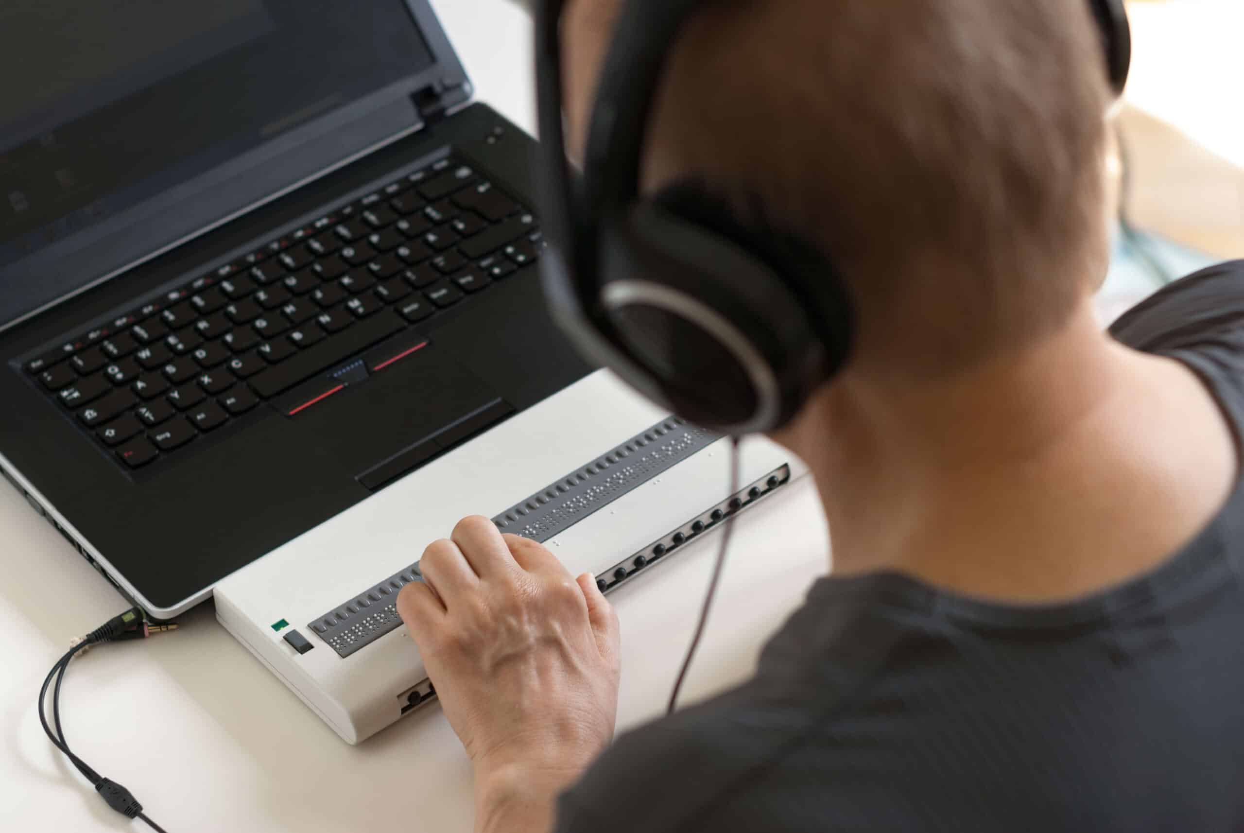 Foto de um homem cego interagindo com um leitor de tela e um display braille, usando fones de ouvido.