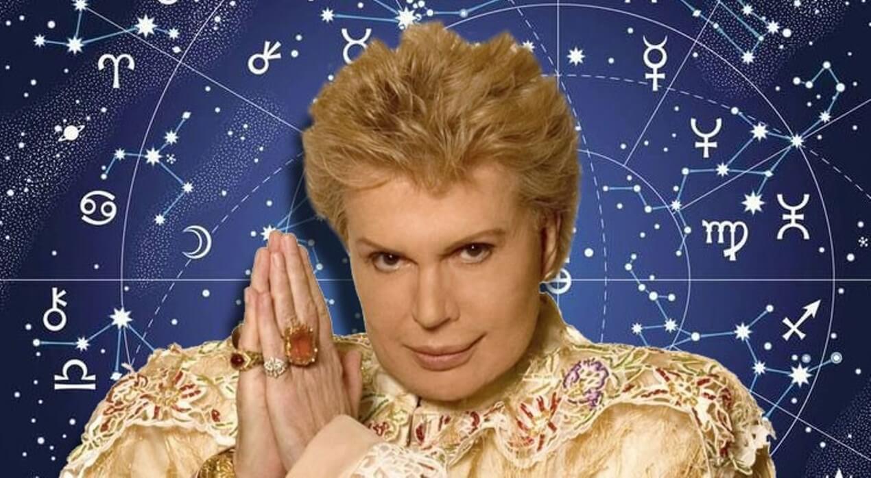 Foto do astrólogo e vidente Walter Mercado. Ele olha diretamente para a câmera, com as mãos em posição de prece. Veste uma roupa em um tom claro, dourado, e usa vários anéis de ouro, um deles com uma grande pedra cor de âmbar. Walter está recortado contra um fundo azul, que é decorado com elementos astrológicos.