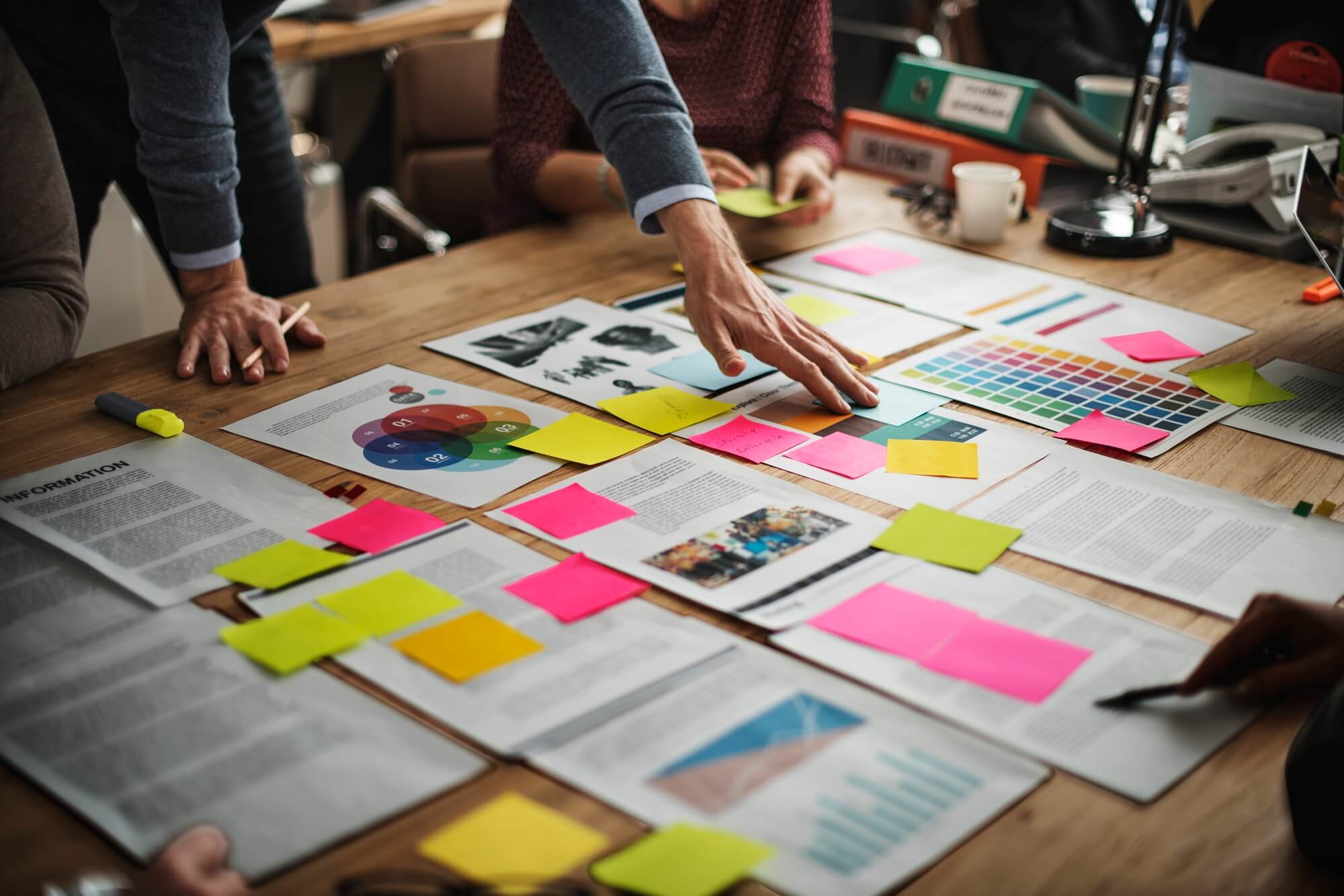 Foto de pessoas em torno de uma mesa de reunião. Sobre a mesa há várias páginas com textos e gráficos, além de post-its coloridos. Uma pessoa está de pé, apontando para uma das páginas.