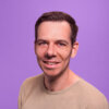 Tim Metz’s headshot. He is standing before a violet background, looking into the camera and slightly smiling.